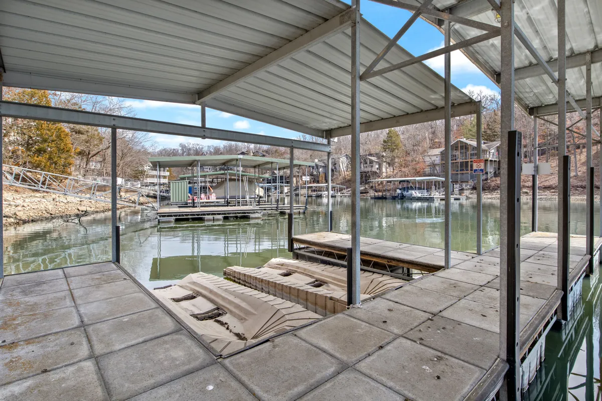 A boat dock stretches over calm water, surrounded by green trees with houses nestled in the background. The scene is lively yet peaceful, with the dock ready for boats, fishing, or relaxing after a day on the lake. The layout suggests space for both boats and personal watercraft.