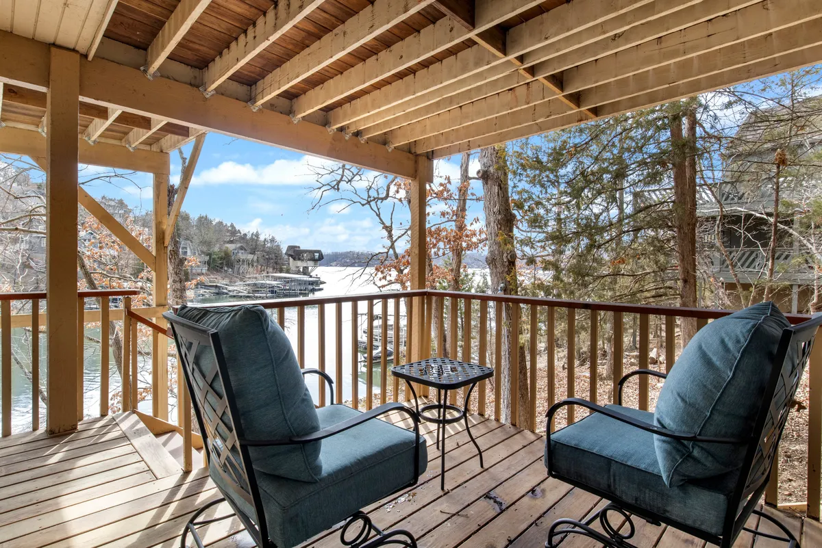 A serene seating area on a lakeside deck, surrounded by mature trees. Comfortable outdoor furniture invites relaxation while offering unobstructed views of the peaceful lake. The setting is secluded and immersed in nature, ideal for quiet mornings or intimate evenings under the stars.