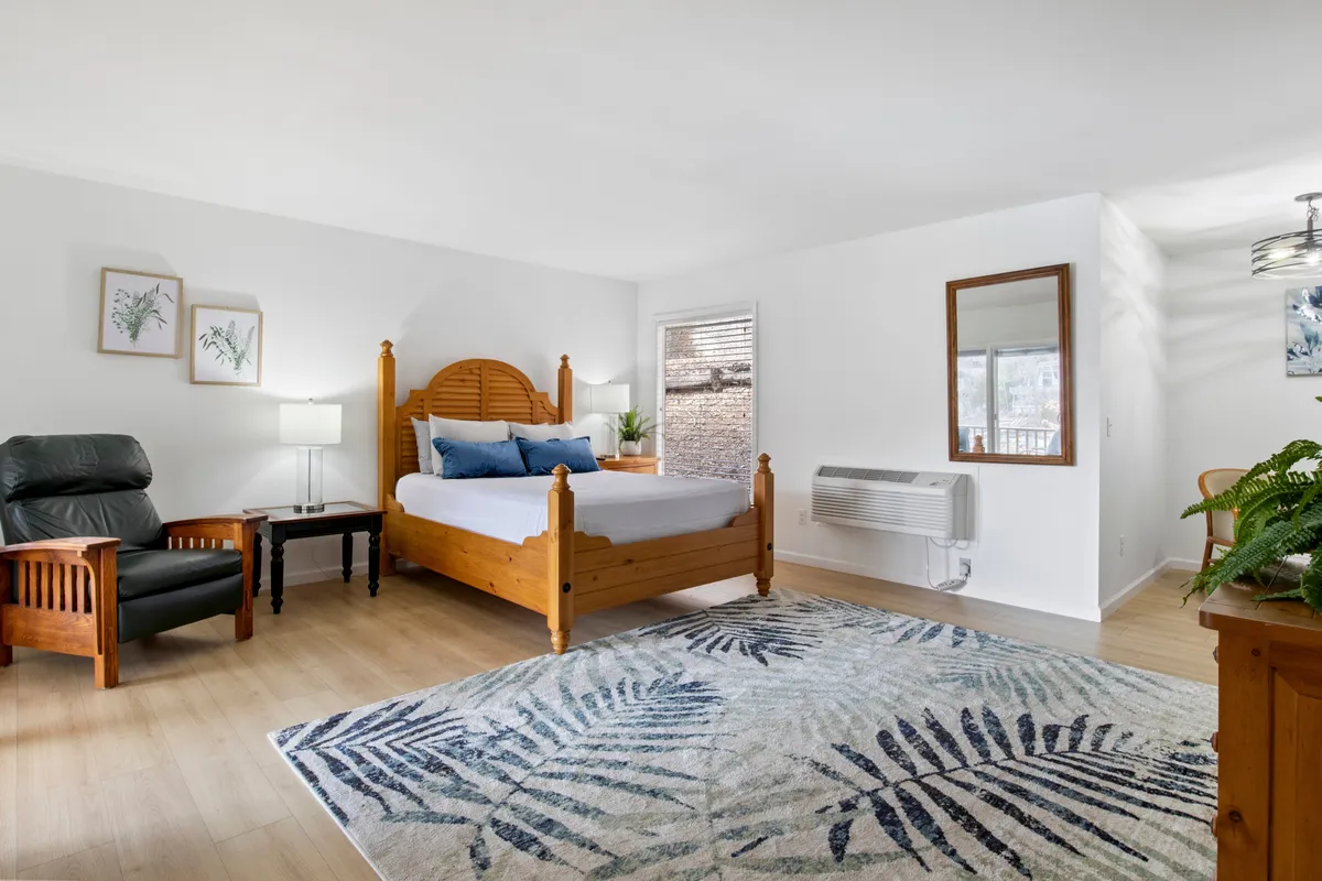 A serene bedroom with a wooden bed frame, soft textured rug, green plant, wooden chair, and gentle lighting from a bedside lamp. The decor is simple and calming, with cozy linens and natural elements.