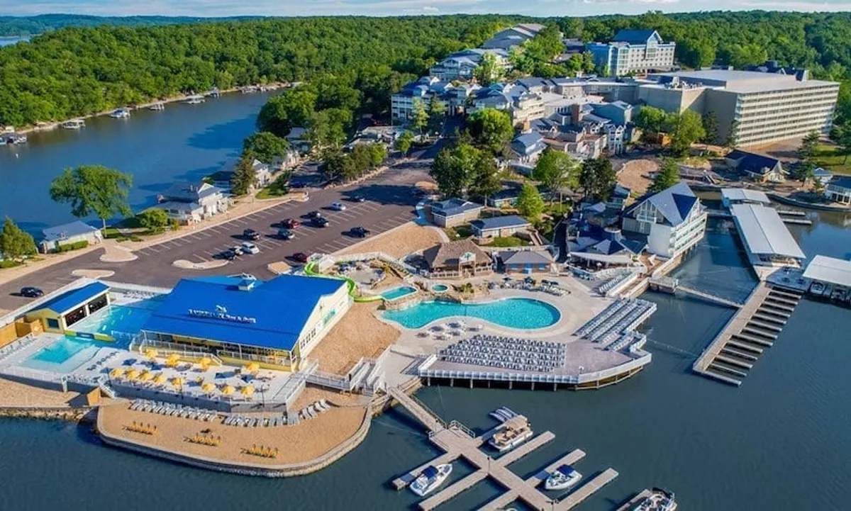 A vibrant lakeside scene featuring a large swimming pool lined with colorful beach chairs, boats docked at a nearby marina, and resort buildings in the background. The area buzzes with activity—swimming, boating, and waterfront relaxation—all under a warm sunset glow.