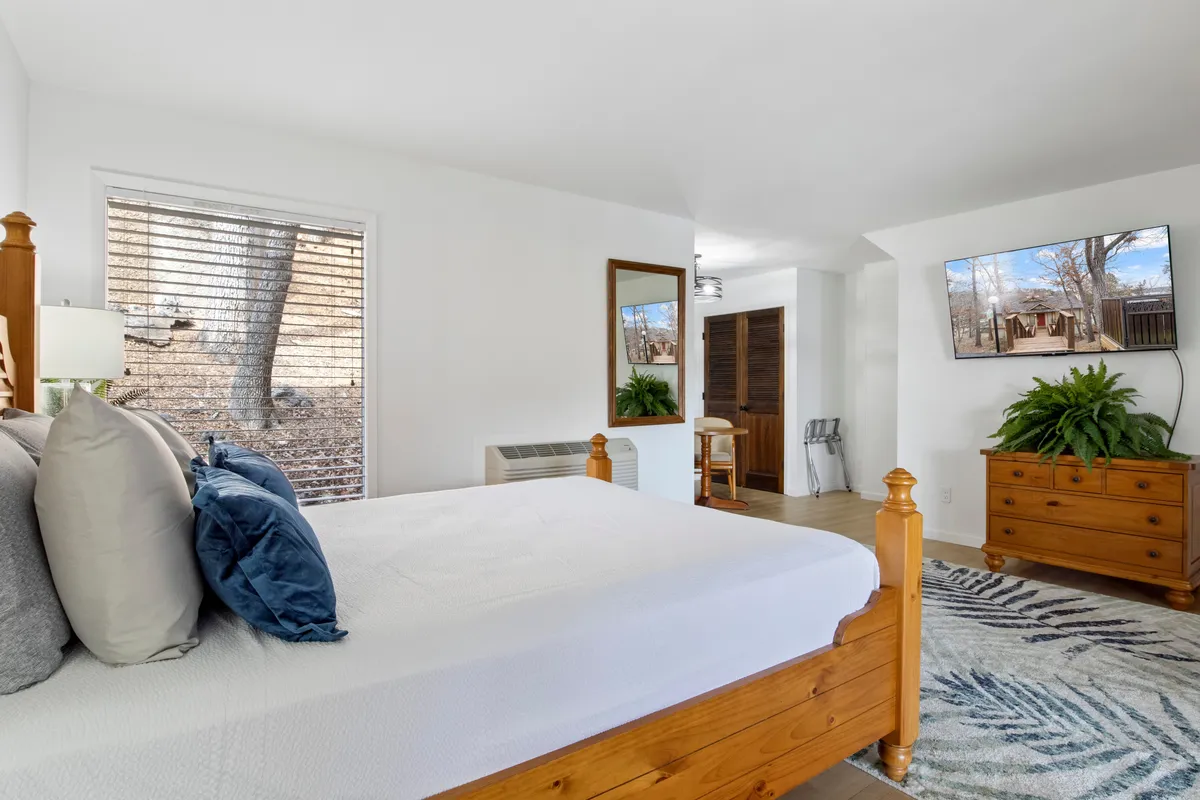 A sunlit bedroom with a large bed adorned with decorative pillows, a wooden dresser topped with a plant, a TV mounted on the wall, and a large window with blinds. The room feels airy, comfortable, and updated.