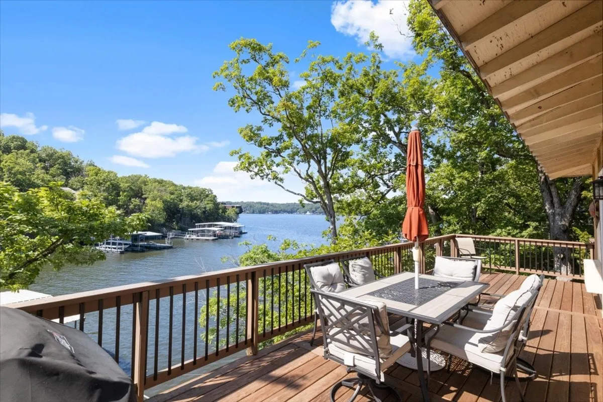 The hero image showcases a spacious outdoor deck with a stylish dining set beneath a vibrant orange umbrella, overlooking serene lake waters framed by lush trees. A private dock with boats is visible, inviting guests for fishing or boating adventures. The tranquil setting is perfect for sunset dinners, morning coffee, or afternoon barbecues, embodying lakeside relaxation and recreation.