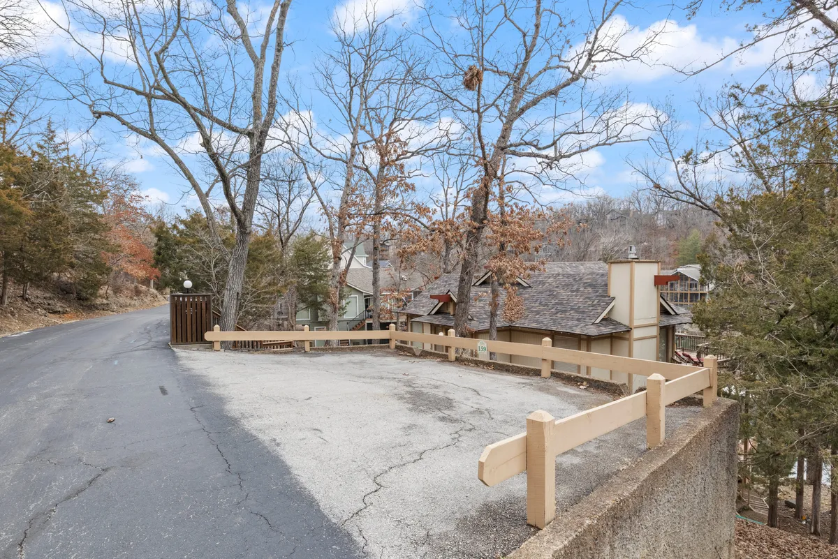 A wide shot of a paved road bordered by a fenced parking area, flanked by trees and residential buildings in the background. The image evokes a sense of peaceful arrival and connection with nature, with hints of autumn foliage and open space for exploration.