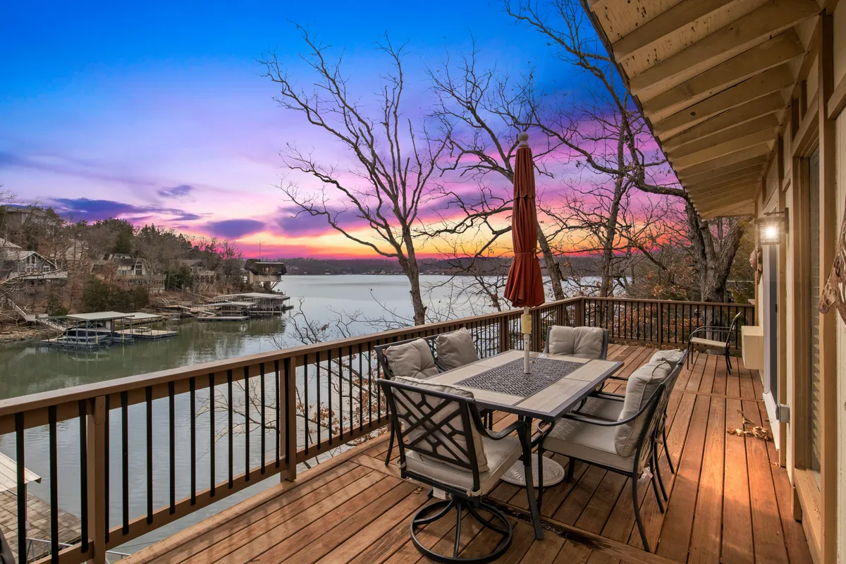 This image presents an outdoor dining table under an umbrella, overlooking a serene lake at sunset. The scene is set for al fresco meals, group celebrations, or morning coffee with picturesque water views and gentle sunset colors.