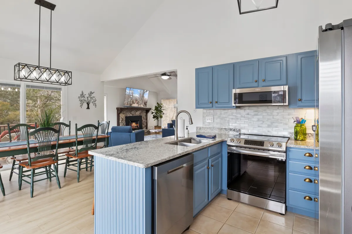 The image highlights a welcoming living and dining area with blue cabinetry, a granite countertop, a dining table, and a fireplace. The open layout seamlessly connects the kitchen, dining, and living zones, capturing the modern finishes and warmth described in the content.
