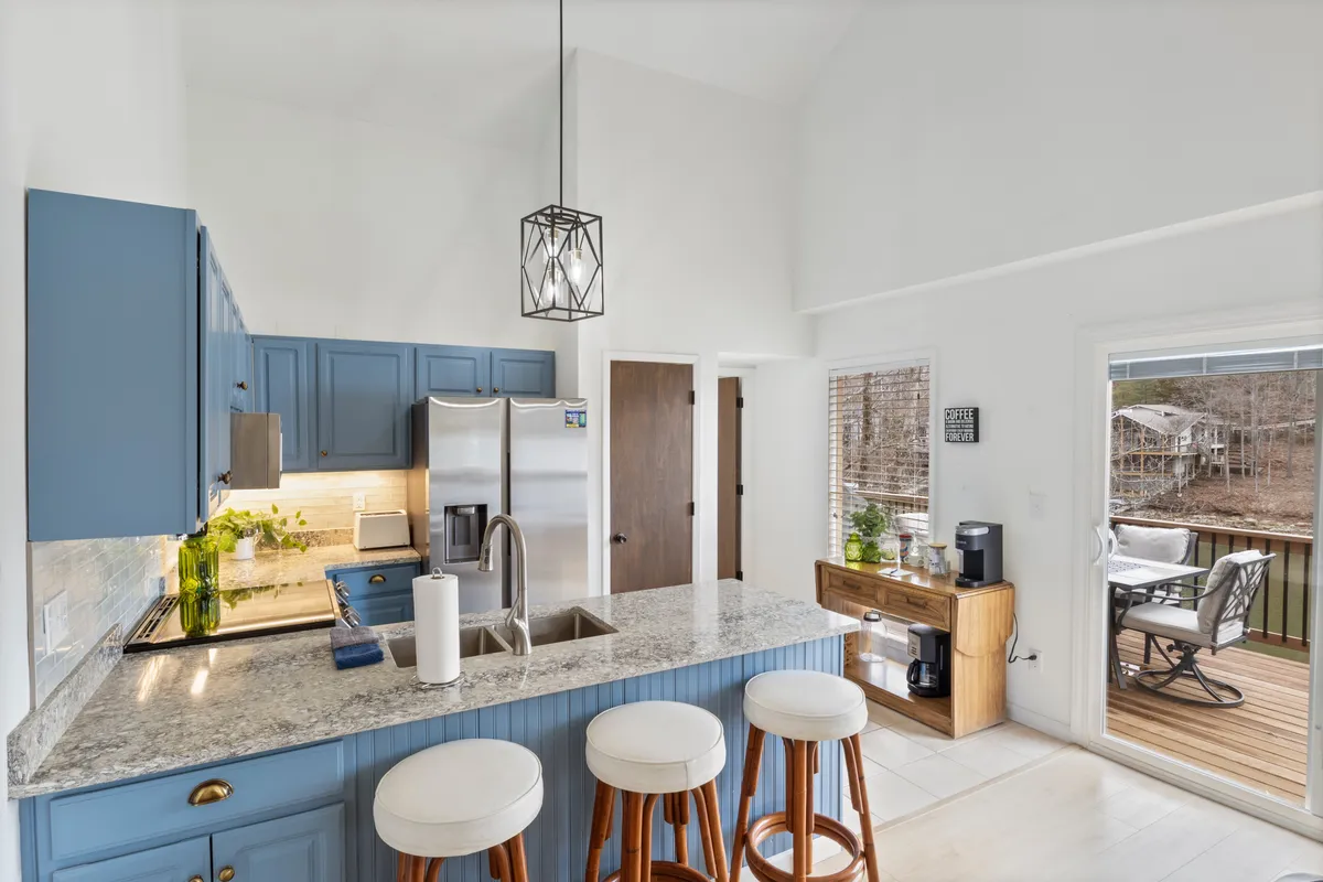 A cozy kitchen with granite countertops, blue cabinetry, pendant lighting, and a view of the outdoor deck through large windows. Stools at the counter suggest a spot for morning coffee or intimate meals while enjoying the view beyond.