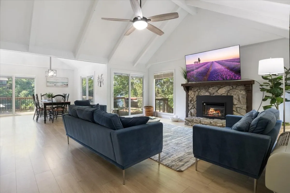 This image features a spacious living room with a sofa, a modern fireplace, a television, and an adjacent dining area. The setting feels open and welcoming, with ample space for family gatherings or cozy evenings. Large windows are visible, allowing natural light to fill the space and offering glimpses toward the outdoors.