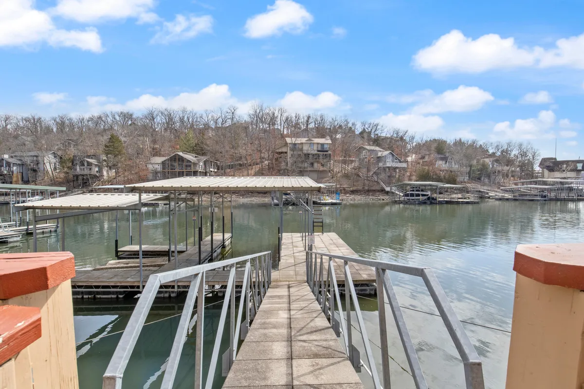 A tranquil dock extends out over the calm waters of Lake of the Ozarks, surrounded by lush trees and nearby lakeside homes. The inviting setting shows ample space on the dock for boats and water activities, with the gentle light hinting at either sunrise or sunset. The water is peaceful, perfect for boating, fishing, or relaxing.